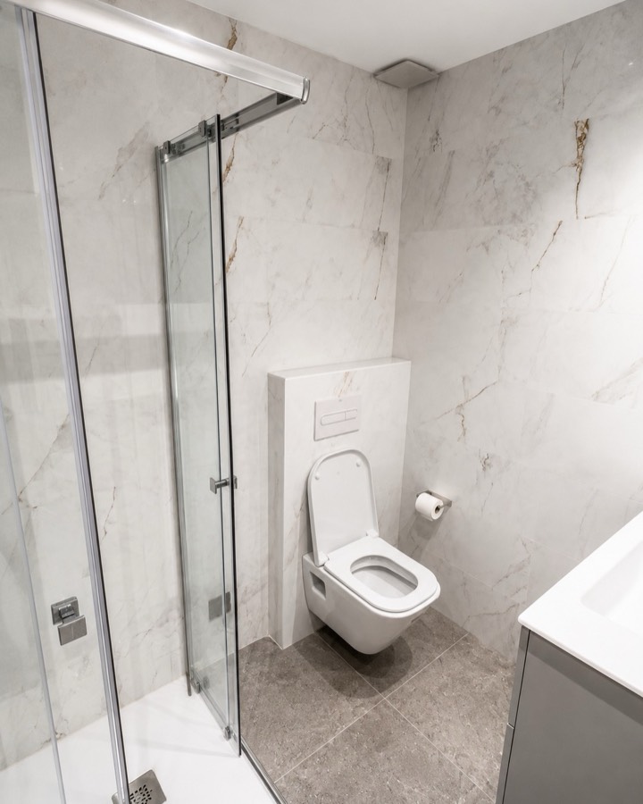 Baño terminado con revestimiento efecto mármol, ducha de cristal e inodoro suspendido.